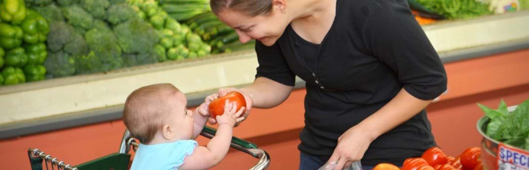 In the fresh produce section of a store, a woman hands a tomato to a baby seated in a shopping cart.