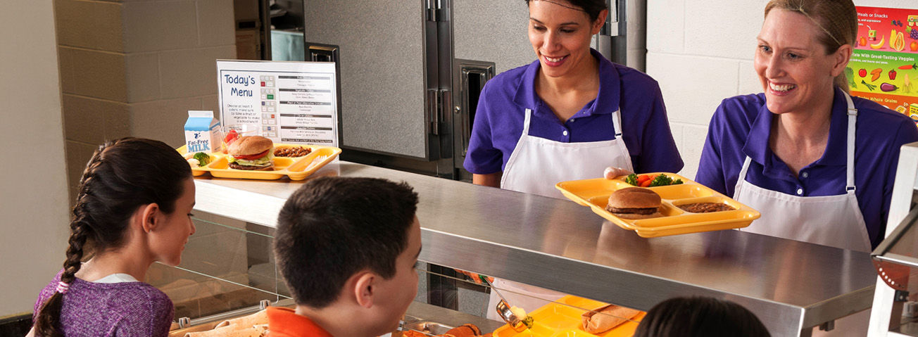 Two female cafeteria workers serve food to several students in a lunch line.