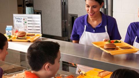 Two female cafeteria workers serve food to several students in a lunch line.